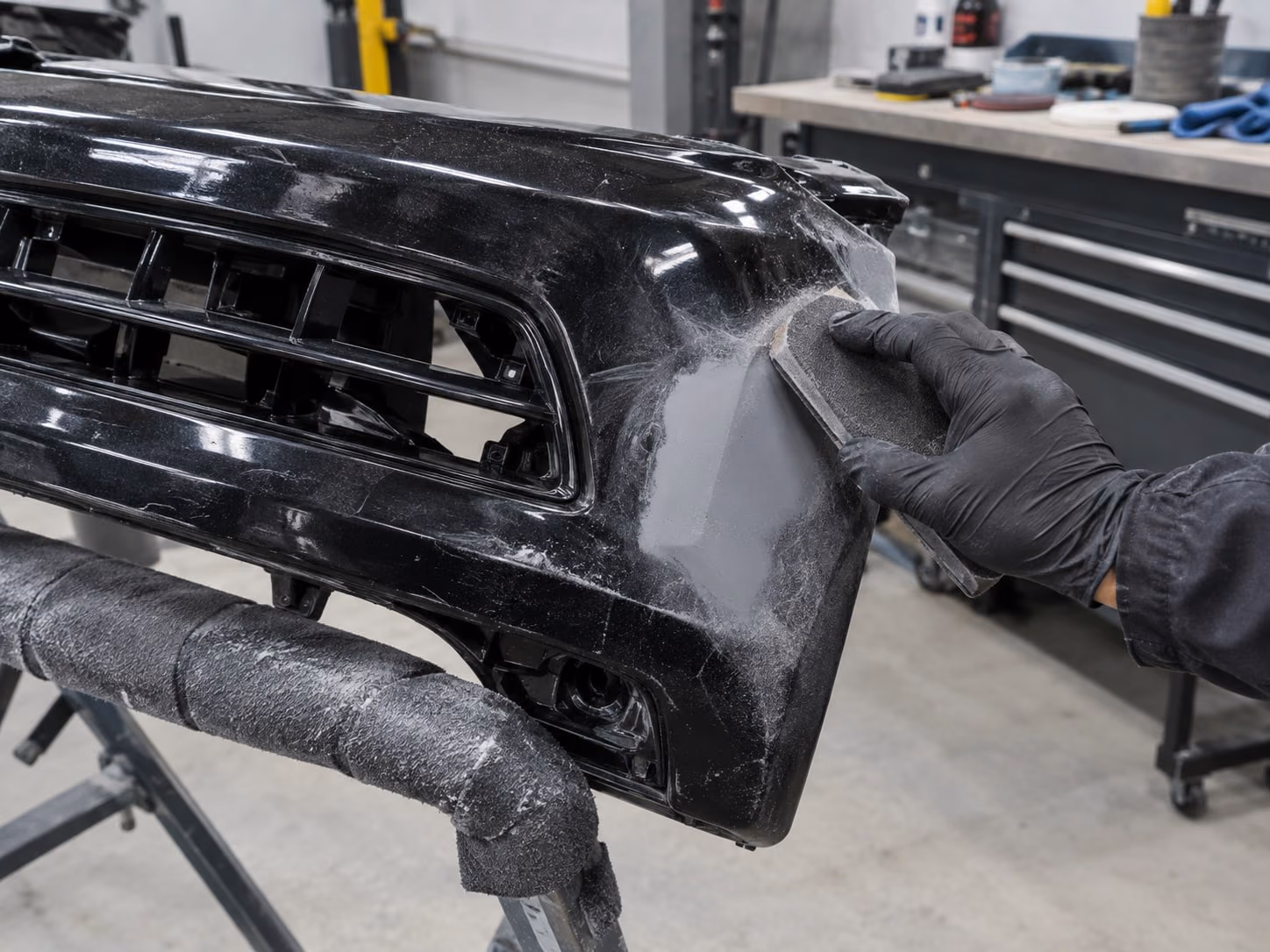 Black front bumper on a padded stand, a gloved hand smoothing a primed corner with a sanding block